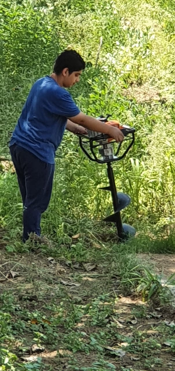 A young man uses a post hole digger to drill a hole at the edge of a clearing.